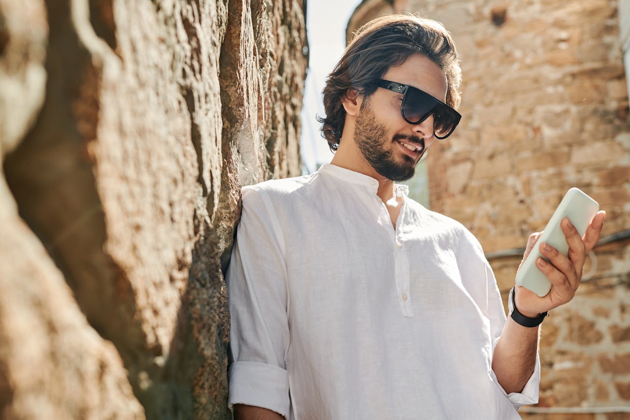 Trendy bearded man enjoying smartphone outdoors by a rustic stone wall.